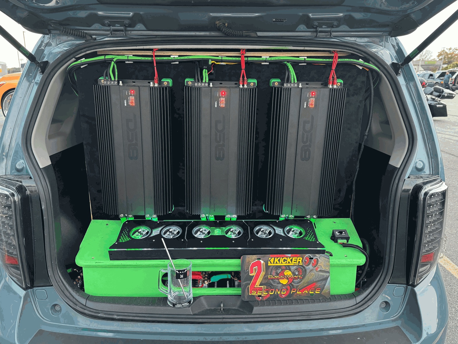 The amp wall in its black and lime finish: three DS18 amplifiers with lit voltage displays over a lime green base, a Kicker Bass Wars second-place plate and a glass second-place mug in front.