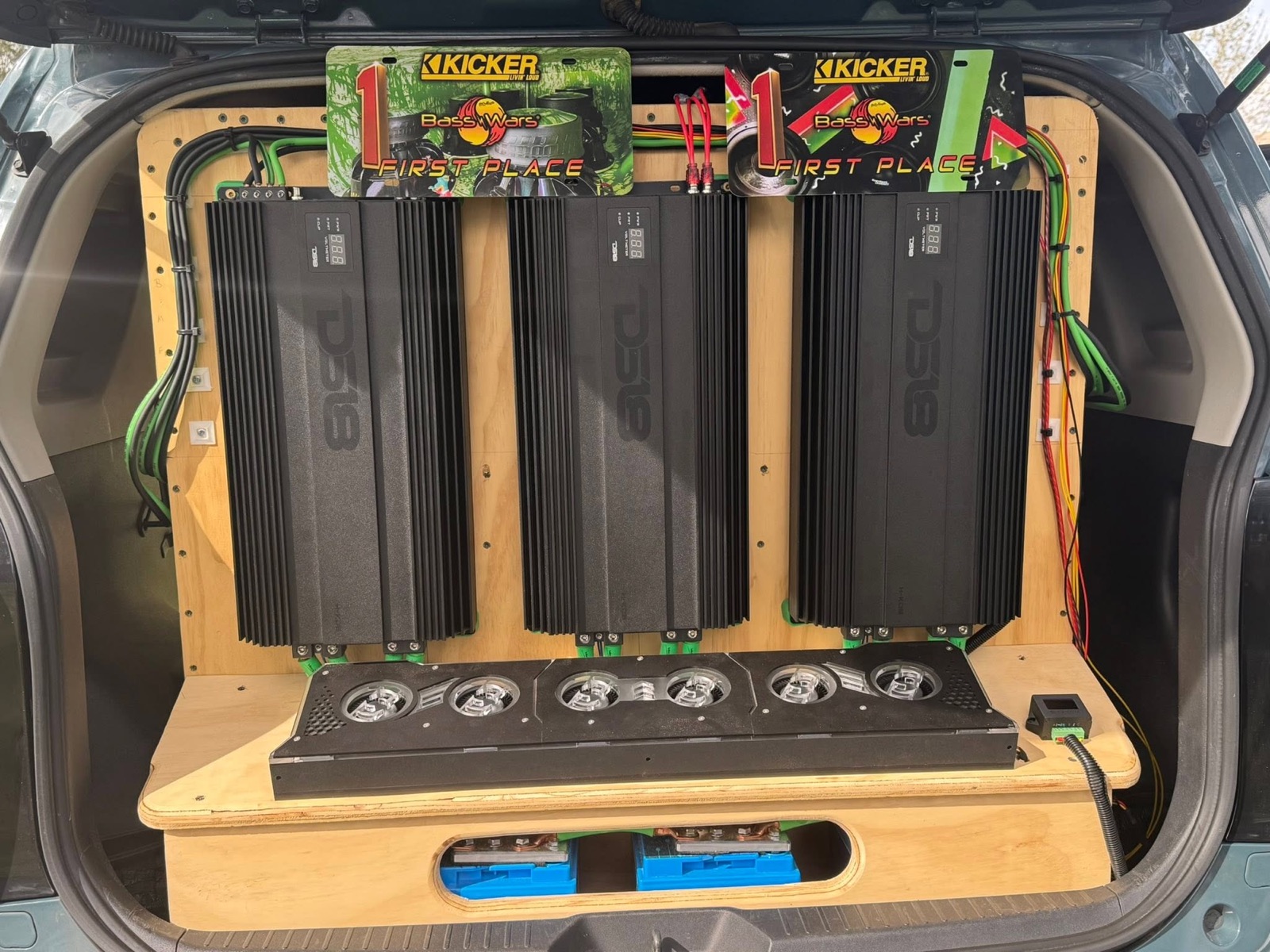 XB Factor's rear hatch open on a plywood amp wall: three black DS18 amplifiers, green and red power cabling, a shelf of cooling fans, a battery bank through a cutout, and two Kicker Bass Wars first-place plates above.