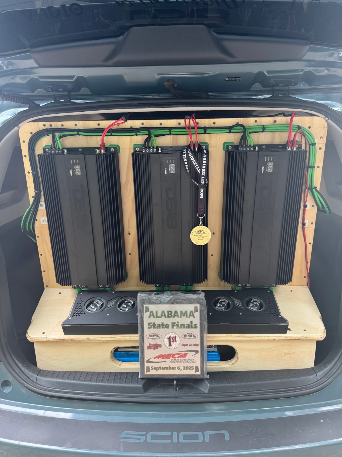 XB Factor's plywood amp wall with a MECA Alabama State Finals first-place plaque dated September 6, 2025, and a MECA SPL Sound Pressure Best of Show medal hanging from the middle amplifier.