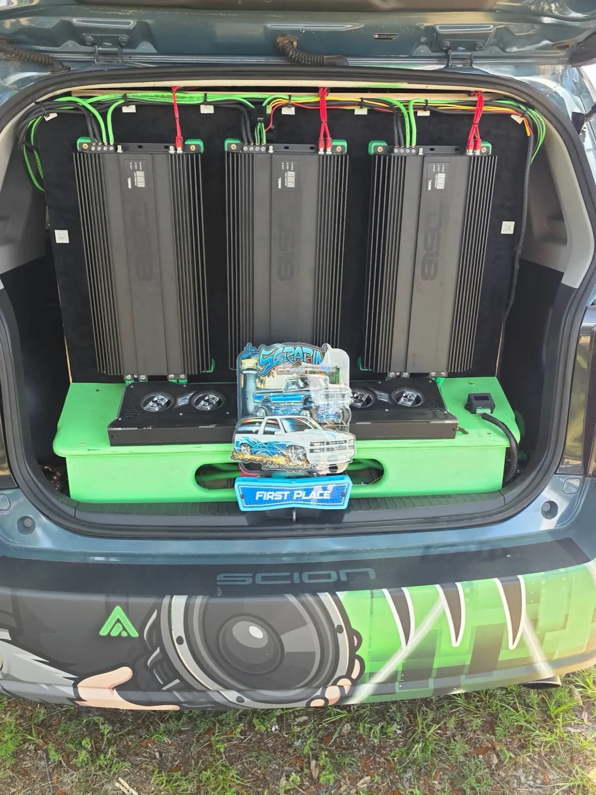 XB Factor's hatch with the amp wall in black and lime, and a Scrapin' the Coast, Biloxi, Mississippi 2026 first-place award in front. The rear bumper wrap shows a hairy hand gripping a subwoofer.