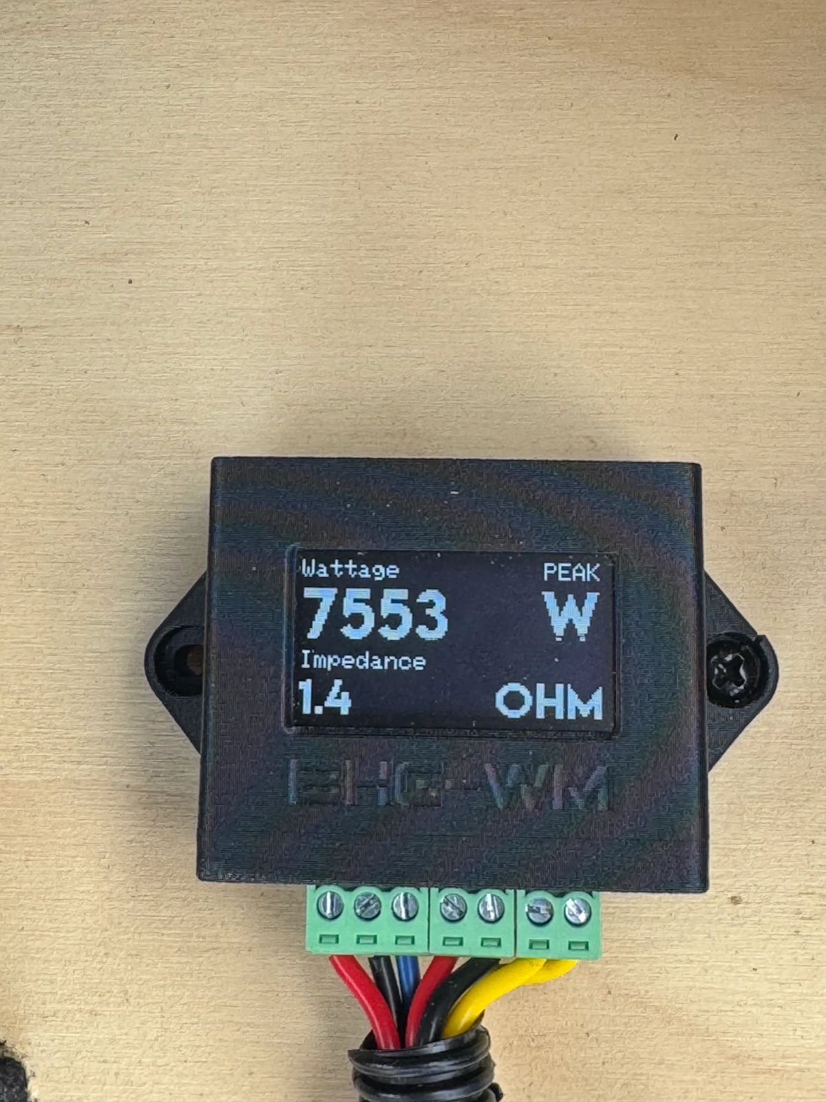 A small BHG-WM watt meter mounted on the plywood wall, showing a peak of 7553 watts at 1.4 ohms impedance.