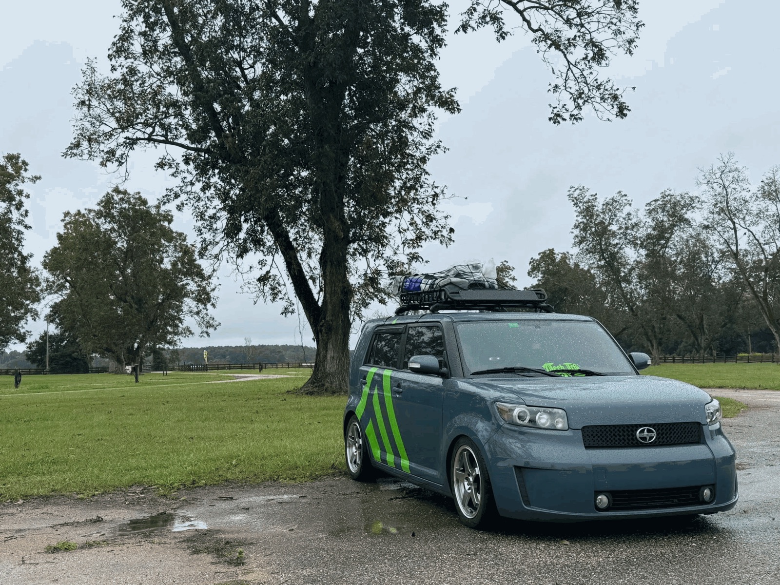XB Factor, a blue-grey Scion xB with lime green chevrons on its doors and a loaded roof rack, parked on a wet gravel drive in front of a large tree.